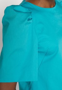 Gros plan d'une personne portant un haut à manches courtes turquoise avec des épaules bouffantes et une texture subtile du tissu.