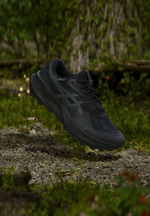 Crna trail running cipela s reljefnim potplatom koja lebdi iznad kamenitog šumskog tla s zelenilom i drvećem u pozadini.