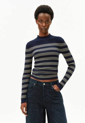 Frau mit kurzen lockigen Haaren, die einen marineblauen, gerippten Pullover mit weißen horizontalen Streifen und dunkle, hoch taillierte Jeans mit Taschen trägt.