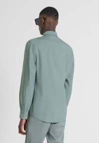 Camicia a maniche lunghe di un verde chiaro, con una texture morbida, polsini con bottoni, vestibilità sartoriale, colletto classico e orlo leggermente curvo nella parte posteriore.