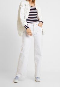 Weiße, weite Jeans mit fransigen Säumen, kombiniert mit einem navy- und weiß gestreiften Oberteil. Eine hellfarbige Jacke rundet das Outfit ab.