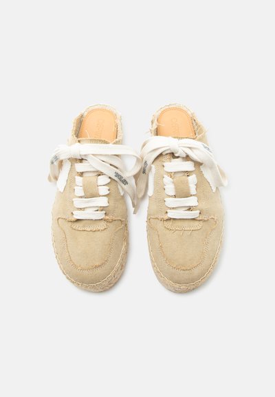 Ζευγάρι μπεζ υφασμάτινων παπουτσιών τύπου slip-on με λευκά κορδόνια δεμένα σε φιόγκους, εμφανείς ραφές και δερμάτινες εσωτερικές σόλες πάνω σε απλό λευκό φόντο.