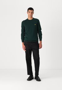 Sweater de malha verde-escuro com decote redondo e mangas longas. Logotipo amarelo no peito. Calças e sapatos pretos completam o conjunto.