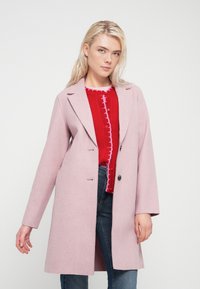 Ροζ παλτό από μείγμα μαλλιού με γιακά με εγκοπές, κλείσιμο με ένα κουμπί και δύο πλαϊνές τσέπες, φορεμένο πάνω από κόκκινο πλεκτό πουλόβερ και μπλε τζιν.