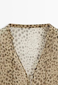 Przezroczysta beżowa bluzka z ciemnobrązowym wzorem w cętki lamparta i dekoltem w szpic z cienkim wiązaniem z przodu.