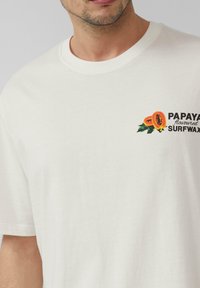 Witte katoenen t-shirt met een kleurrafische afbeelding van papaja en de tekst "PAPAYA smaak SURFWAX" op de linkerkant van de borst. Korte mouwen.