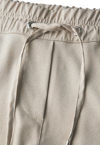 Joggers beige in tessuto texturizzato con vita elasticizzata, chiusura con cordoncino e dettagli in occhioletto metallico. Pieghe funzionali migliorano il design.
