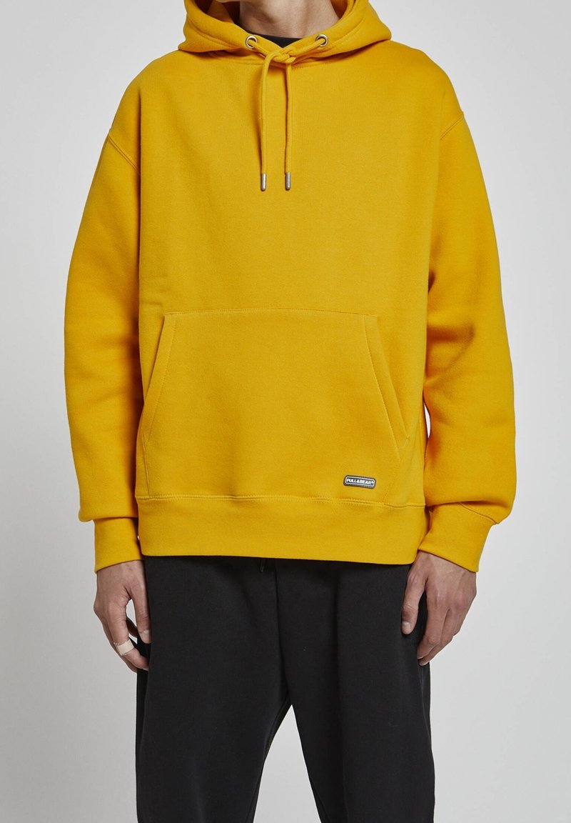 Personne portant un sweat à capuche jaune moutarde avec poche ventrale et pantalon noir, debout devant un fond gris uni.