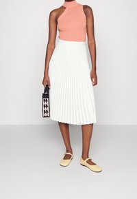 Top sin mangas melocotón, falda midi plisada blanca, bailarinas amarillas y un bolso de mano con patrón en negro y beige. Diseño simple y moderno con texturas suaves.