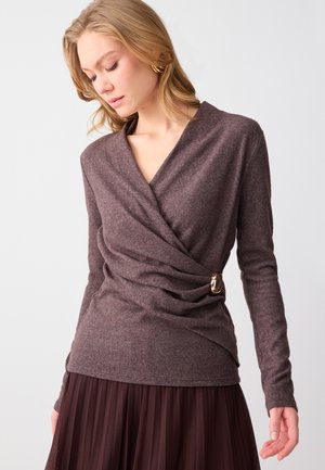 Donna che indossa un maglione incrociato prugna a maniche lunghe con fibbia dorata abbinato a una gonna scura plissettata su sfondo neutro.