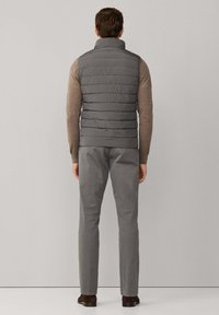 Grijze donsbodywarmer met een hoge kraag, gedragen over een beige shirt met lange mouwen, gecombineerd met grijze broek en donkerbruine schoenen. Geribbelde textuur zichtbaar.