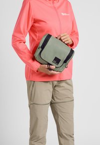 Borsa compatta verde con cerniera nera e logo, tenuta in una mano. La persona indossa una giacca corallo e pantaloni beige. Design semplice e funzionale.