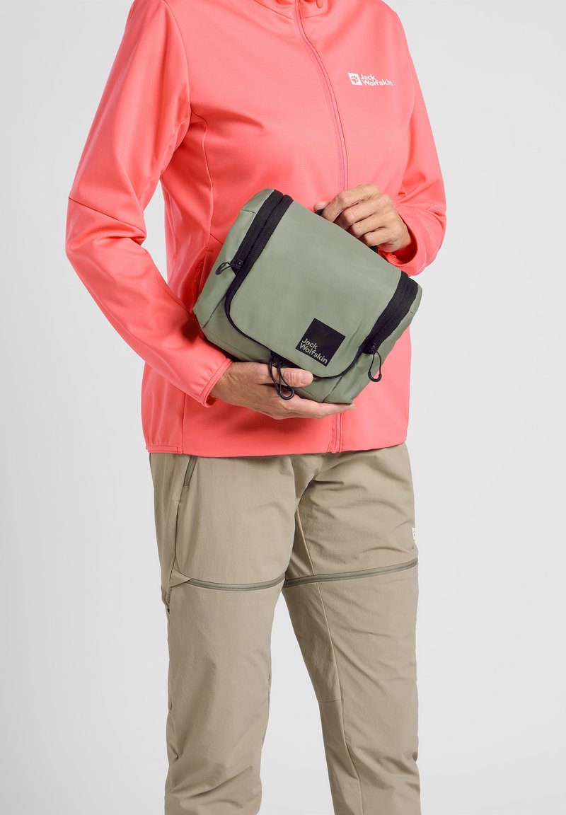 Borsa compatta verde con cerniera nera e logo, tenuta in una mano. La persona indossa una giacca corallo e pantaloni beige. Design semplice e funzionale.