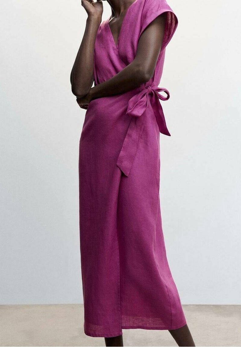 Personne portant une robe portefeuille magenta sans manches, longueur au genou, avec un nœud noué sur le côté, debout devant un fond clair uni.