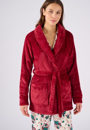 Accappatoio rosso in velour con colletto a scialle, cintura coordinata e tasche frontali. Abbinato a pantaloni da pigiama con motivo floreale. Texture morbida.