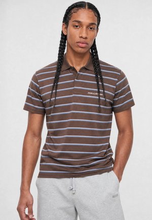 Ung mand med langt fletningehår iført en brun og blåstribet poloshirt og lysegrå joggingbukser med den ene hånd i lommen.