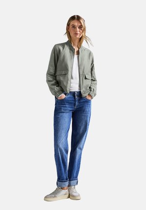 Jeune femme debout les mains dans les poches, portant une veste bomber gris clair, une chemise blanche, un jean bleu retroussé et des baskets beiges.