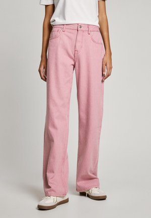 Vrouw draagt roze-wit gestreepte rechte broek met verticaal patroon en zakken aan de voorkant, gecombineerd met witte sneakers en een wit T-shirt.