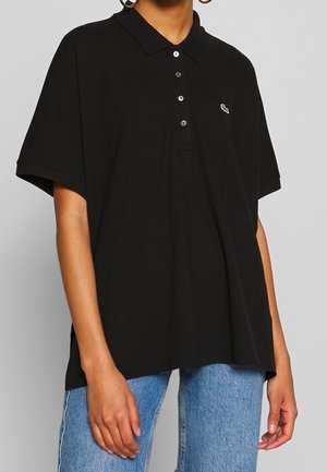 Osoba ubrana w luźną czarną koszulkę typu polo z czterema guzikami i małym logo krokodyla, ubrana w jasnoniebieskie dżinsy.