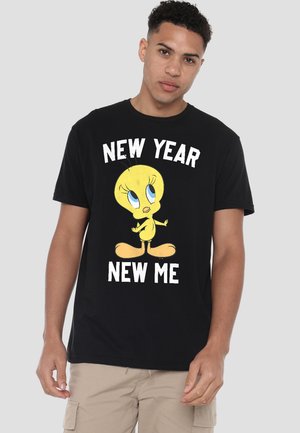 Young man wearing black T-shirt with Tweety bird graphic and text "New Year New Me," paired with beige cargo shorts, standing against gray background.