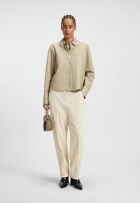 Frau mit geflochtenem Haar, die ein beige knopffrontiertes Hemd und creamfarbene weite Hose trägt, hält eine taupefarbene Handtasche und steht vor weißem Hintergrund.