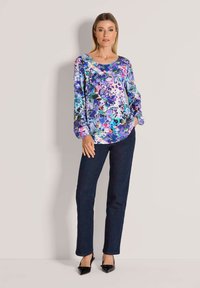 Floralbluse in Lila-, Blau- und Pinktönen mit langen Ärmeln und rundem Ausschnitt, kombiniert mit dunkelblauen Jeans und schwarzen High Heels.