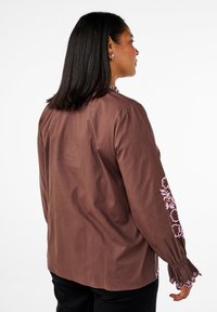 Blusa marrone a maniche lunghe con ricamo floreale rosa sulle maniche e polsini a scallop. Realizzata in tessuto morbido con una vestibilità comoda.
