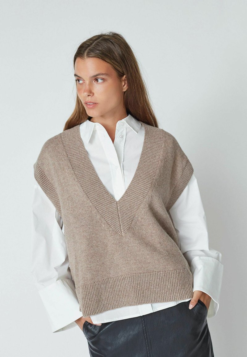 Light brown knitted vest with a deep V-neck, short sleeves, ribbed hem, worn over a white collared shirt, set against a neutral background.