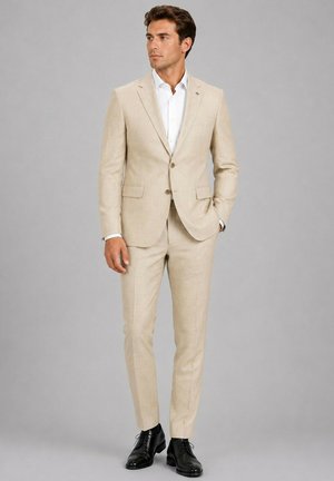 Homme portant un costume beige avec une chemise blanche et des chaussures habillées noires, debout devant un fond gris clair uni.