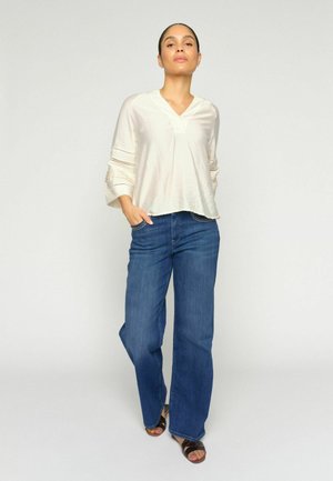 Žena stojaca s rukami v kapsách, oblečená v krémovej blúzke, modrých širokých džínsoch a čiernych sandáloch, na neutrálnom pozadí.