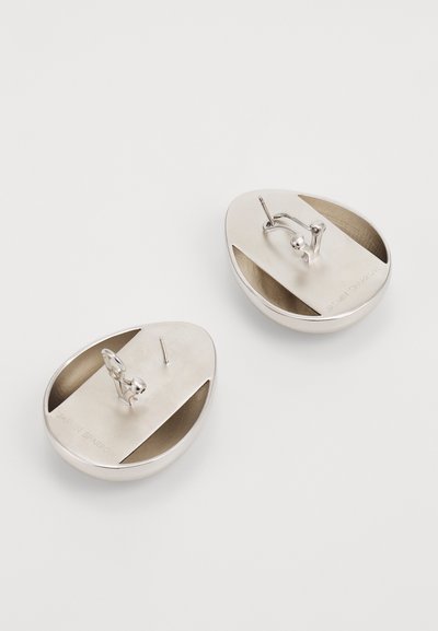 Silver earrings with an oval shape, featuring a smooth textured surface and cut-out sections. The back has a fixed hook and pin for fastening.