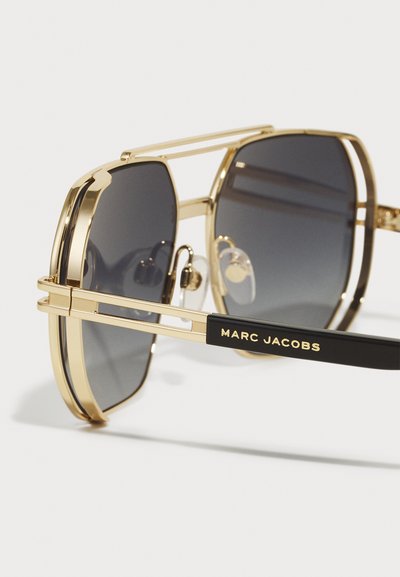 Auksiniai metaliniai saulės akiniai su juodais įdėklais. Yra dideli tamsūs objektyvai, dviguba tiltelio forma, ir išgraviruotas "MARC JACOBS" logotipas ant šono.