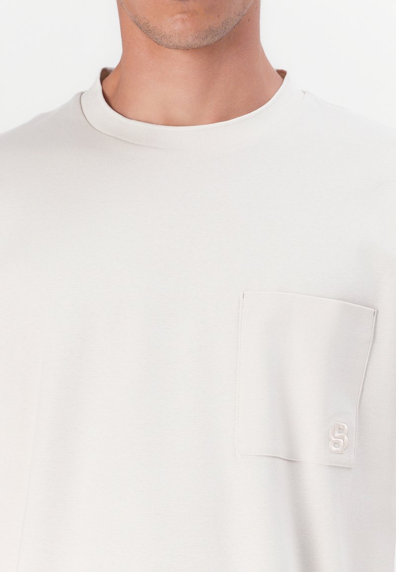 Jasnoszara bawełniana koszulka z okrągłym dekoltem, krótkimi rękawami oraz kieszonką na piersi z subtelnym haftem logo. Gładka tekstura.