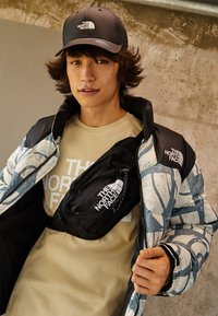 Giacca trapuntata blu e bianca con motivo, camicia beige con testo, borsa nera con logo, cappellino grigio. Tessuto morbido e colori a contrasto.