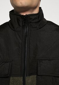 Zwarte zip-up jas met hoge kraag, textuurstof, twee voork微信 pockets en een contrasterend olijfgroen paneel aan de onderkant.