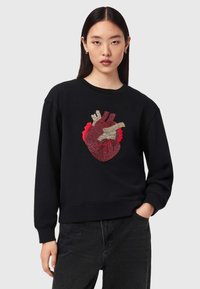 Νεαρή γυναίκα με ίσια μαύρα μαλλιά, φορώντας μαύρο φούτερ με λεπτομερή σχέδιο κόκκινης και μπεζ ανατομικής καρδιάς.