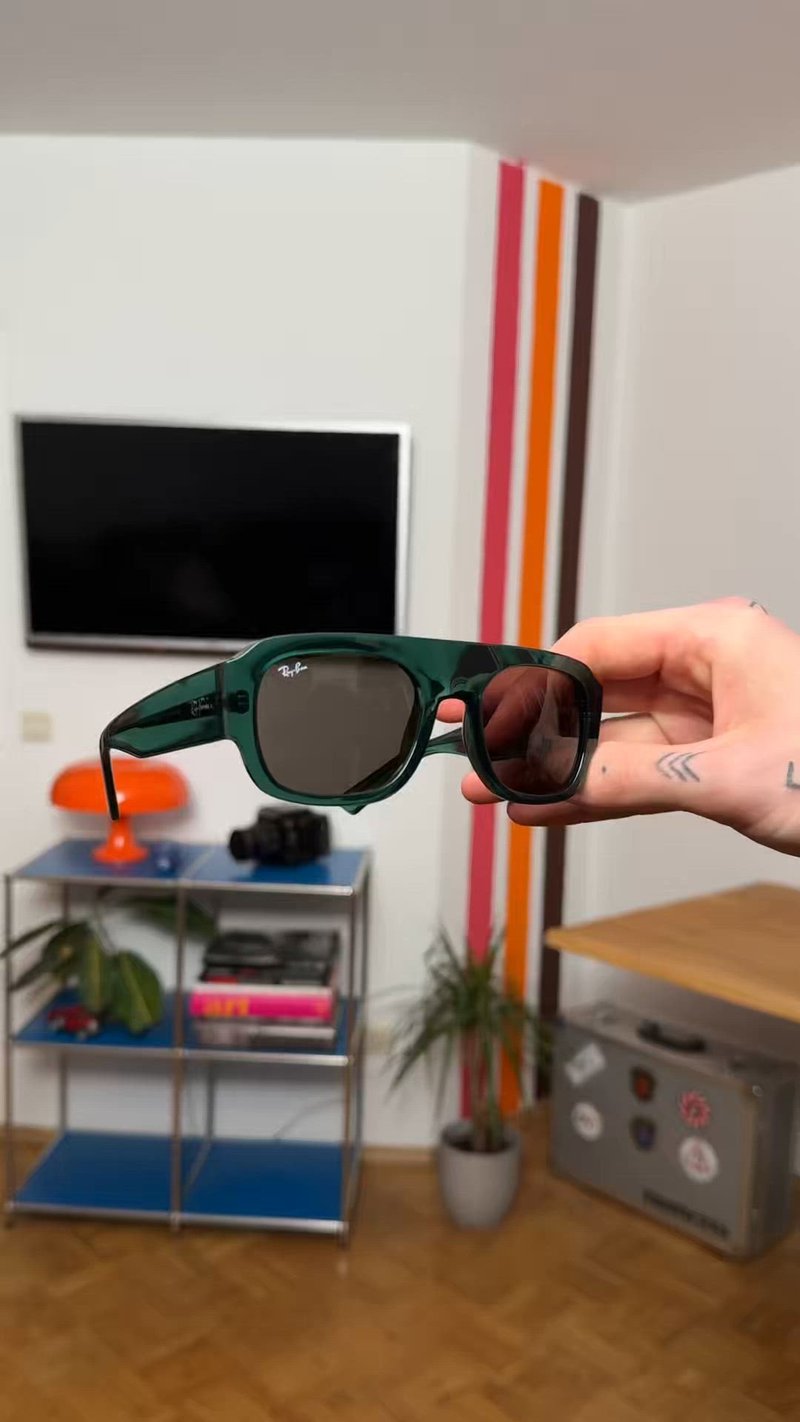 Ръка държи зелени полупрозрачни слънчеви очила Ray-Ban в стая с телевизор, оранжева лампа, метални рафтове, домашни растения и ивици на стената.