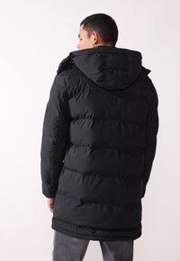 Chaqueta puffer negra con capucha, con textura acolchada, detalles con cremallera y diseño hasta la rodilla. La vista trasera muestra un tejido suave y una silueta elegante.