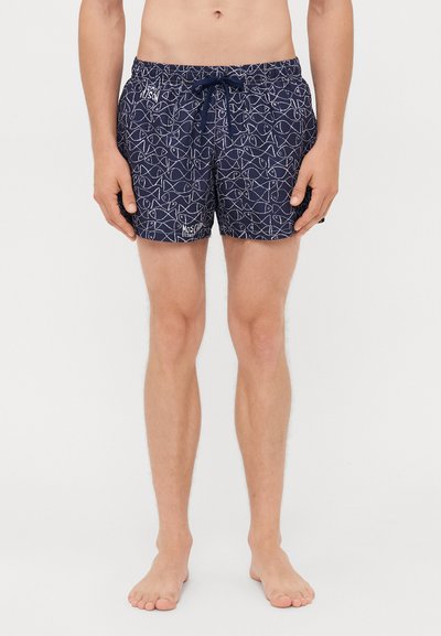Man wearing navy blue swim shorts with white continuous fish line pattern and drawstring waist, standing barefoot against a white background.