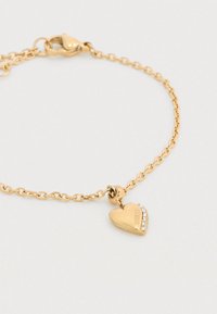 Βραχιόλι με αλυσίδα από χρυσό που περιλαμβάνει μενταγιόν σε σχήμα καρδιάς με σειρά από κρύσταλλα. Η καρδιά είναι γυαλισμένη με χαραγμένη επωνυμία. Κλείσιμο με κόπιτσα.