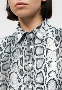 Γυναίκα με πουκάμισο κουμπωμένο μέχρι πάνω με ασπρόμαυρο αφηρημένο σχέδιο φιδιού και κοντά σκούρα μαλλιά.