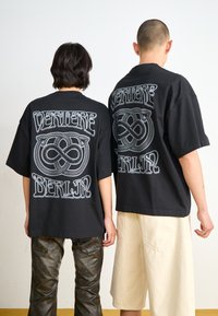 Black oversized t-shirts featuring a silver graphic print with the words "VERTERE" and "BERLIN" and an infinity symbol, worn by two individuals.