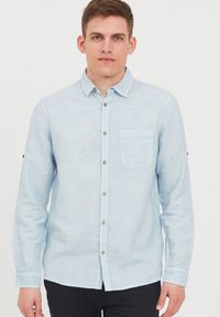 Camisa de manga larga azul claro hecha de una tela texturizada. Presenta un cierre frontal con botones, un bolsillo en el pecho y botones marrones en la tapeta.