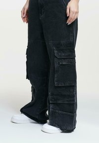 Black cargo pants with multiple pockets, a loose fit, and contrasting stitching, paired with white sneakers on a light background.