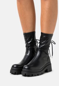 Black leather ankle boots with gathered design, adjustable ties, and chunky treaded sole. Smooth texture with a rounded toe and thick heel.