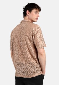 Camicetta a maniche corte in tessuto marrone chiaro con un intricato motivo a diamante con ritagli, che mostra texture e un design strutturato.