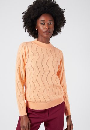 Peach-colored sweater with a textured wave pattern, long sleeves, a ribbed crew neckline, and a fitted hem. Worn with burgundy pants.
