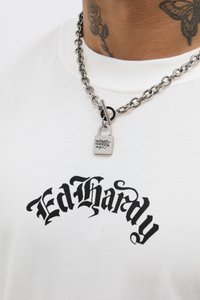 Bijela majica s crnim natpisom "Ed Hardy" u stiliziranom fontu. Metalna ogrlica s malim privjeskom u obliku brava.