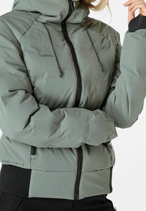 Femme portant une veste matelassée légère grise à capuche avec des fermetures éclair noires et une taille côtelée, les bras croisés, ajustant le cordon de la capuche.