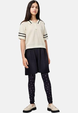 Jeune femme portant un pull crème à manches courtes avec des rayures noires, un short noir, des leggings noirs avec des motifs de cœurs, et des mocassins beige, debout.